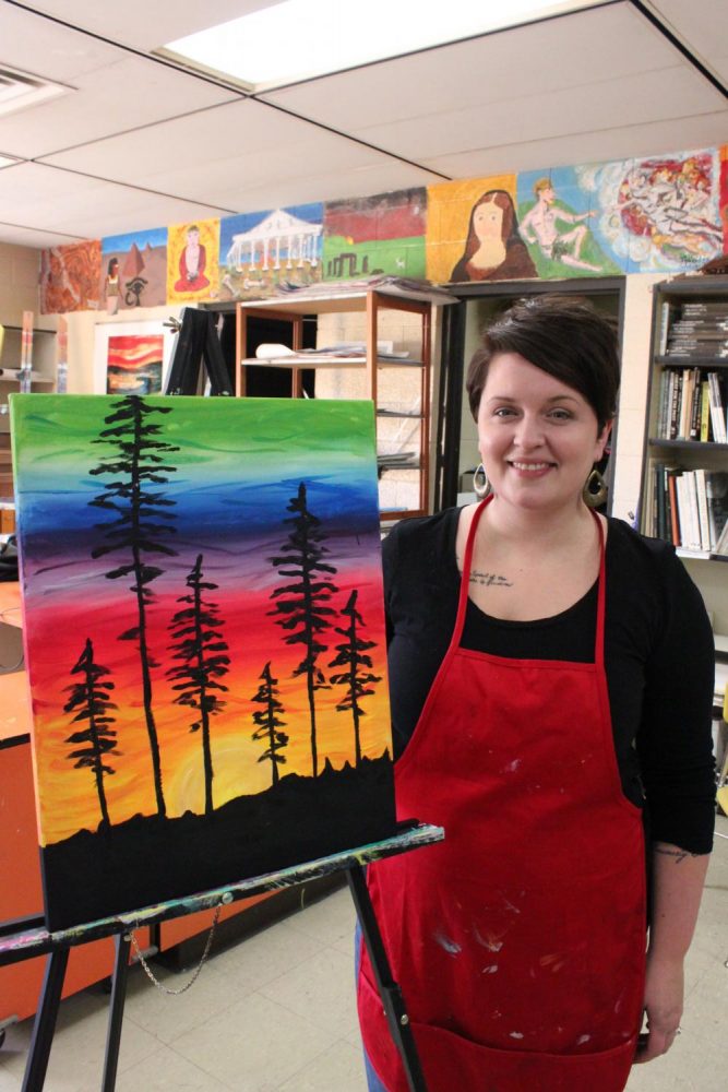 Art teacher Elisha Penning proudly stands by her finished painting after teaching the six students to paint the picture of a colorful sunset with trees for the background.